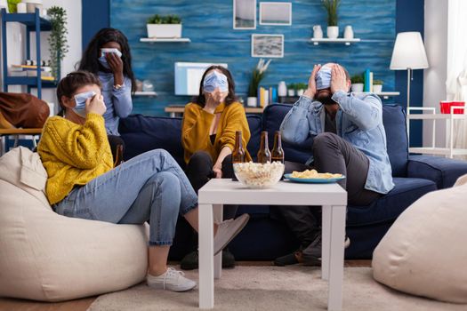 Multicultural friends intimidated looking at scary movie during coronavirus pandemic wearing face mask to prevent illness covering faces sitting on sofa living room.