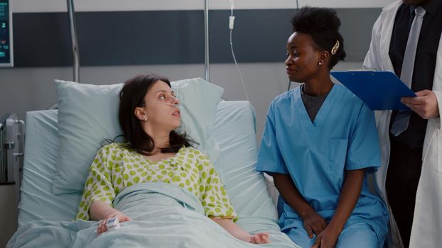 Patient sitting in bed while asisstant with black skin examining disease symptom writing medical expertise on clipboard. Black nurse discussing illness therapy working in hospital ward