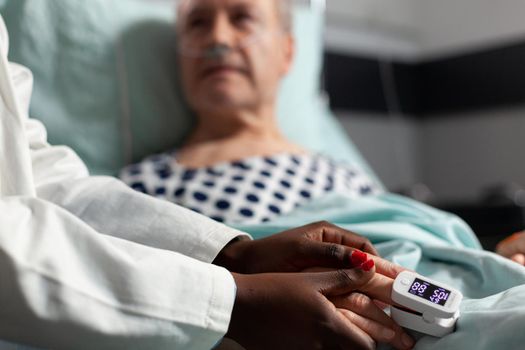 Friendly doctor reassuring sick old patient holding hands, showing emphaty, encouragement, during medical examination in hospital room. Patient breathing through oxygen mask