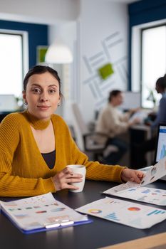 Businesswoman working with company charts looking at camera holding cup of coffee. Executive entrepreneur, manager leader sitting working on projects with diverse colleagues.