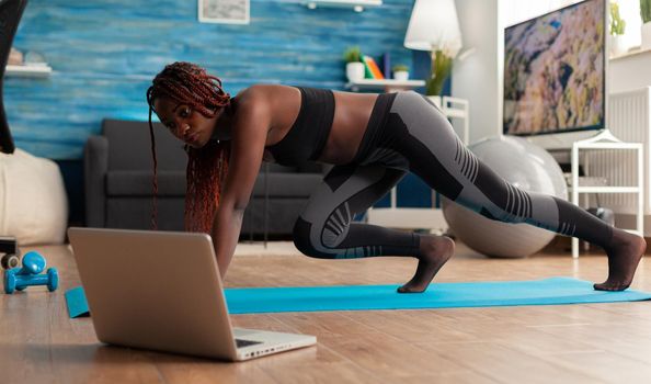 Fit strong black woman doing running plank on fitness mat on living room floor, practicing muntaing climbers workout style for healthy lifestyle wearing leggings and black top.