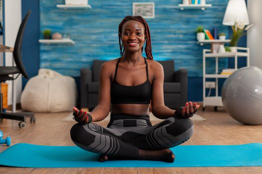 Fit black woman practicing yoga sitting with legs crossed in sportive top and leggins on lotus pose. Practicing calm mind harmony for no stress life in home living room.