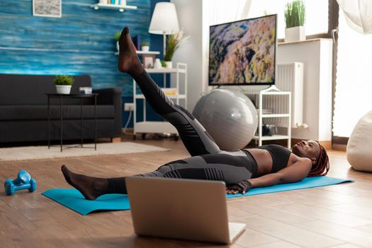 Active strong fit muscular fit woman on exercise mat doing scissors in home living room for legs and abs muscle, in home living room following program from laptop.