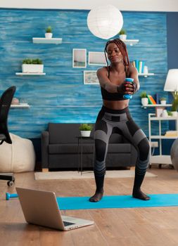 African athlete doing squats with dumbbell dressed in sport wear while watching online class tutorial on laptop during workout at home staying on yoga mat, for strong legs and healthy lifestyle.