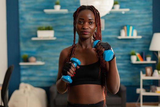 Black woman taking care of body working training arm muscles using dumbbells weights during workout. Positive joyful sportive strong fit athlete in home living room.