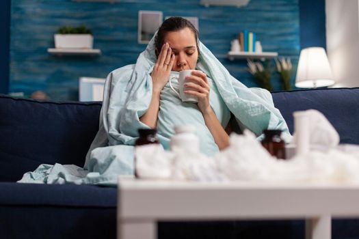 Sick woman sitting at home in blanket with hot tea suffering from fever temperature infection flu headache cold. Young person drinking tea, with seasonal virus symptoms feeling unwell