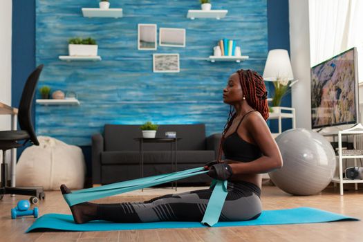 Fit strong afro woman training home living room using resistance band, sitting on fitness mat pulling for back muscles. Training using arm rowing movement holding rubber band.