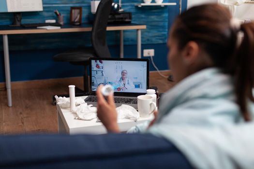 Caucasian woman on webcam communication with doctor for medications support against flu cold virus fever. Young patient at home using telemedicine for healthcare, holding a thermometer