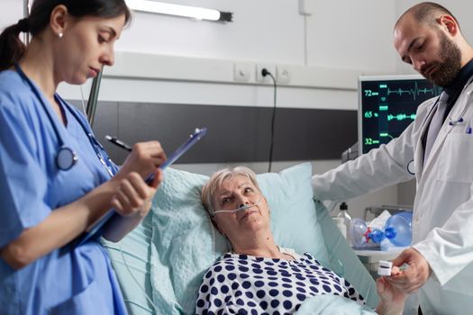 Sick senior woman looking at medical staff laying in hospital bed with oxygen mask to help her inhale and exhale. Doctor checking oxygen saturation and blood pressure on oxymeter attached on finger.