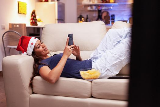 Woman lying on sofa browsing on social media using smartphone during christmastime in xmas decorated kitchen. Girl enjoying winter holiday celebrating christmas season at home