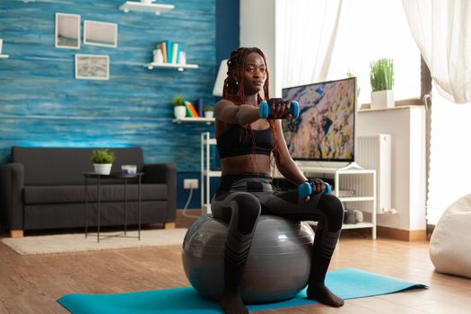 African woman using stability ball keeping outstretched arms working out shoulders using blue dumbbells, in home living room for muscle shaping and healthy lifestyle, dressed in sportwear.