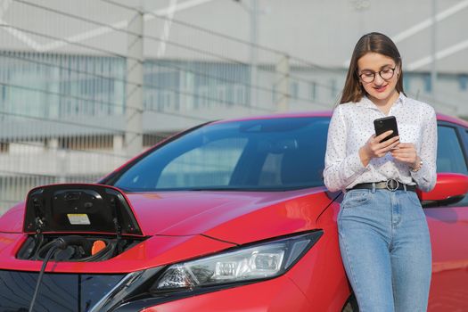 Ecological car connected and charging batteries. Girl using smartphone and waiting power supply connect to electric vehicles for charging the battery in car.