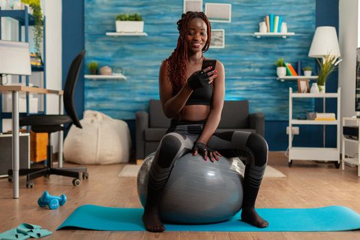 Active athletic black woman chatting on smartphone sitting on swiss ball in home living room, after working out on yoga mat to get stronger body and healthy lifestyle.