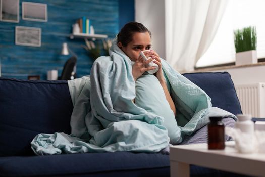 Sick woman at home resting with cup of tea on sofa wrapped in blanket. Unwell young adult with cold flu disease infection fever temperature headache covid 19 virus symptoms