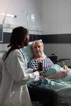African american therapist in hospital room discussing treatment holding pills bottle reassuring recovery,sick senior man laying in bed breathing through oxygen mask,