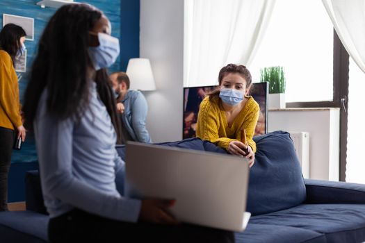 Happy cheerful woman holding beer bottle looking at laptop talking with african friend in apartment living room wearing face mask to prevent coronavirus spread in time of global pandemic. Conceptual image.