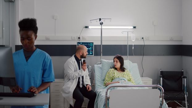 Physician medic consulting sick patient during medical appointment in hospital ward. Doctor discussing medication treatment explaining pill prescription while sick patient resting in bed