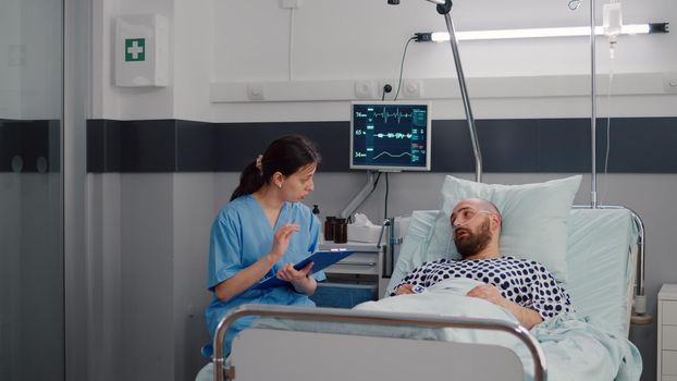 Medical nurse discussing illness treatment with hospitalized sick man resting in bed during rehabilitation therapy in hospital ward. Patient with nasal oxygen tube explaining disease symptom