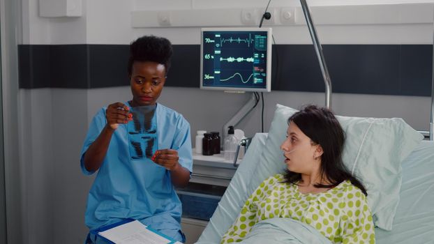 Closeup of nurse with black skin analyzing bones radiography monitoring healdcare recovery. Sick woman patient resting in bed discussing disease treatment in hospital ward during therapy consultation