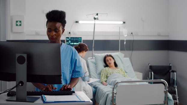 Black assistant typing healdcare treatment on computer while in background surgeon doctors checking sick woman writing illness diagnosis. Patient lying in hospital ward bed during consultation recovery