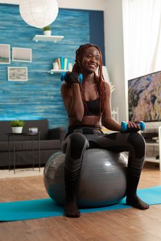 Smiling cheerful african woman flexing arm working out biceps, using dumbbells sitting on stability ball making training harder after warm up. Strong athletic person doing sports at home.