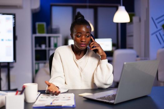 Black businesswoman manager discussing important project during phone call. Busy focused freelancer doing overtime for job reading writing, searching, respecting deadline of financial project, overworking.