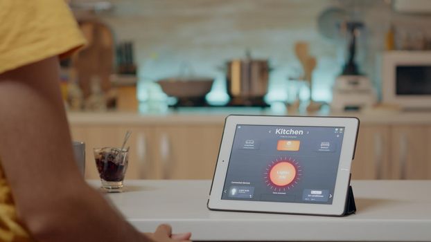 Man looking at tablet with wireless lighting automation software placed on kitchen desk, house with smart system, turning on lights. Digital tablet with high tech app controlling electricity effiency