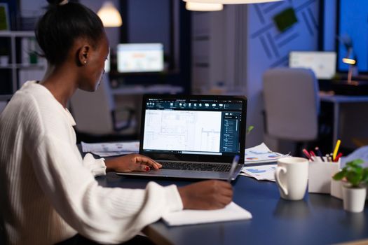 African architect working late at night on laptop with blue prints, looking at graph. Industrial woman employee studying prototype idea showing cad software on device display.