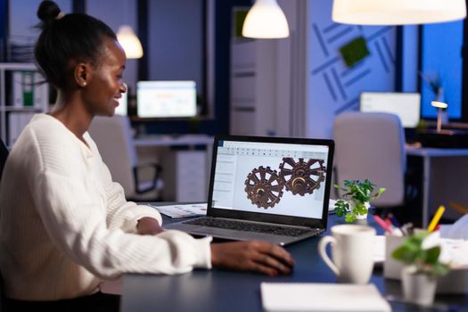 African engineer working late at night on a 3D model of industrial gears at laptop doing overtime in start up office. Freelancer studying prototype idea on pc showing cad software on device display.