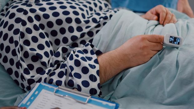 Closeup of sick man patient resting in bed with medical oximeter on finger. Surgeon doctor checking heart pluse analyzing illness cardiogram working in hospital ward explaining disease diagnosis