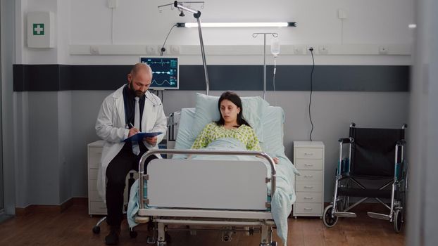 Front view of sick woman resting in bed discussing with oncologist man doctor about test results. Practitioner writing sickness treatment on clipboard monitoring healdcare recovery in hospital ward