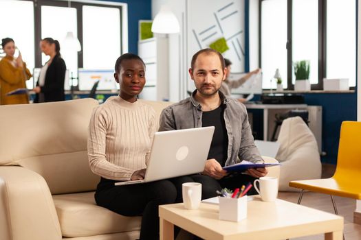 POV of diverse colleagues discussing about financial strategy during video meeting using webcam sitting on couch in start up office, working for new business. Multiethnic coworkers analysing reports.