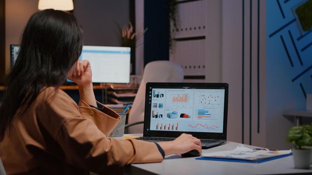 Overworked businesswoman typing financial statistics on laptop sitting at desk table in startup company office late at night. Workaholic entrepreneur planning economic project before company deadline