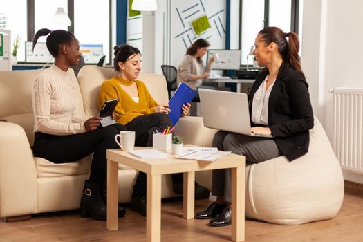 Group of multiethnic business people discussing about company strategy sitting on coach in modern cooperation office looking at statistics. African entrepreneur having a meeting with investors.