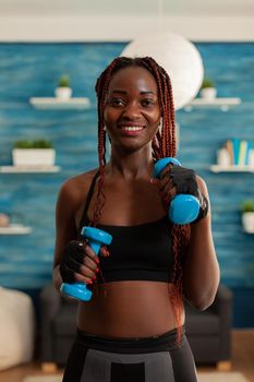 Fit black woman exercising biceps with dumbbells in home living room, dressed in sportwear. Sportive strong active african smiling at camera during workout with healthy lifestyle.