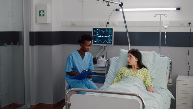 Sick woman resting in bed while afro-american nurse monitoring disease writing medical healthcare treatment on clipboard. Assistant discussing illness therapy working in hospital ward