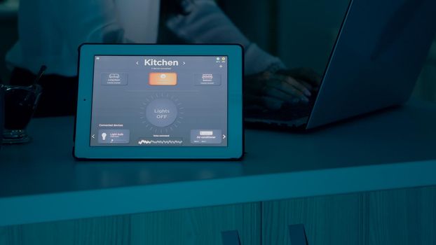 Female typing on laptop working from home with automation lighting system using voice controlled on tablet switching on bulbs. Smart gadget responds to commands, woman using automated app software