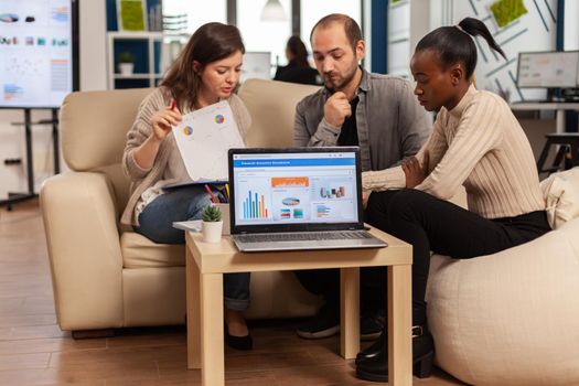 Diverse colleagues employees cooperating in office discussing at workplace, analysing graphs sitting on couch. Business coworkers planning start up business project, brainstorming in corporate office