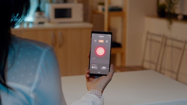 Female holding smartphone with lighting control application, turning on the lights sitting in kitchen house with automation lighting system. Woman using high technology software, wireless switch on bulbs