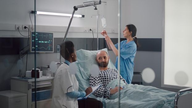 Medical team monitoring patient vital checking heart rate injecting fluids in drip bag for remission therapy in hospital ward. Sick woman waiting for healthcare treatment during clinic examination