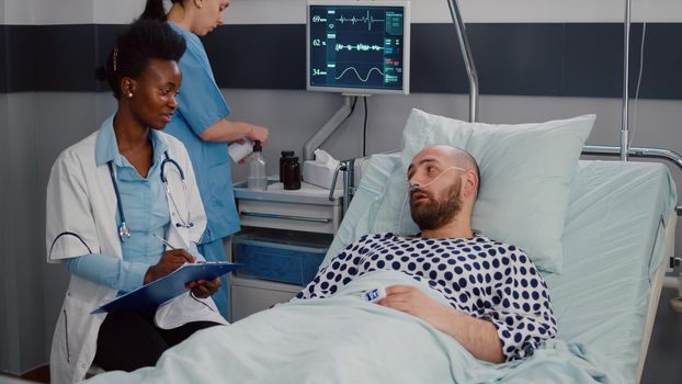 Afro american doctor discussing with sick man writing medical treatment on clipboard in hospital ward during illness rehabilitation. Patient resting in bad with nasal oxygen tube in therapy room