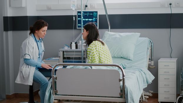 Sick woman adult sitting in bed while practitioner examining disease expertise writing medical sickness treatment on clipboard. Doctor discussing illness therapy working in hospital ward