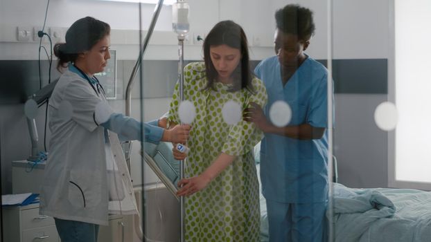 Medical team helping sick woman to stand up from bed holding intravenous IV fluid drip bag after disease surgery in hospital ward. Practitioner checking expertise recovery examining illness symptom