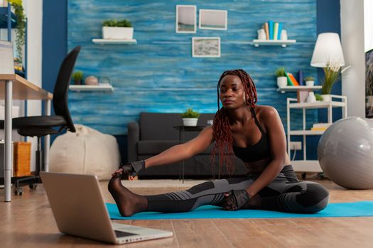 Sporty fit black woman doing stretching flexibility exercise reaching toes sitting on fitness mat in home living room watching tutorial video on laptop screen. Workout and healthy lifestyle.