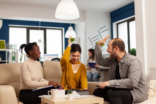 Diverse team of successful young entrepreneurs sharing high-five celebrating success sitting on couch in start up office room. Multiethnic business team with laptop and papers excited about project