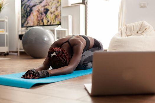 Black woman exercising and stretching her body doing flexibility exercises sitting on yoga mat in home living room wearing leggings during online tutorial. Workout and healthy lifestyle.