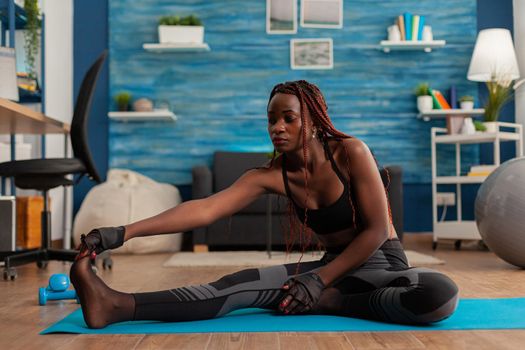 Fit black woman stretching reaching toes sitting on yoga mat after intense workout in home with dumbbells training. Relaxing muscles for healthy lifestyle wellbeing.