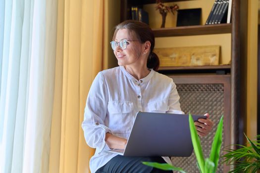 Middle-aged woman psychologist in an office with laptop. Portrait of professional female counselor, psychiatrist, therapist, teacher sitting on chair near window. Technology, medicine, people