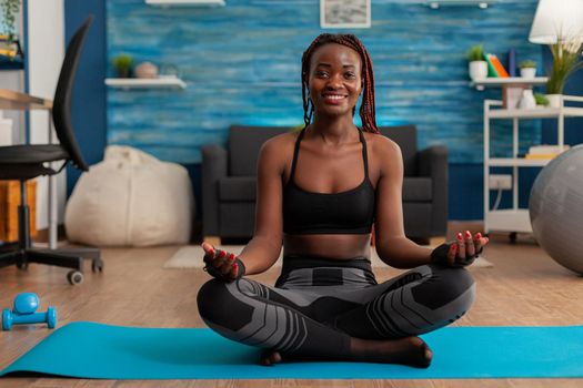 Fit black woman practicing yoga sitting with legs crossed in sportive top and leggins on lotus pose. Practicing calm mind harmony for no stress life in home living room.