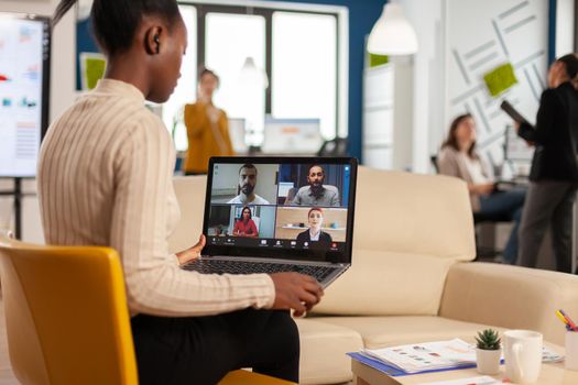 African manager woman discussing with remote colleagues using video call holding laptop sitting on couch in business modern office. Diverse coworkers planning new financial project in modern company